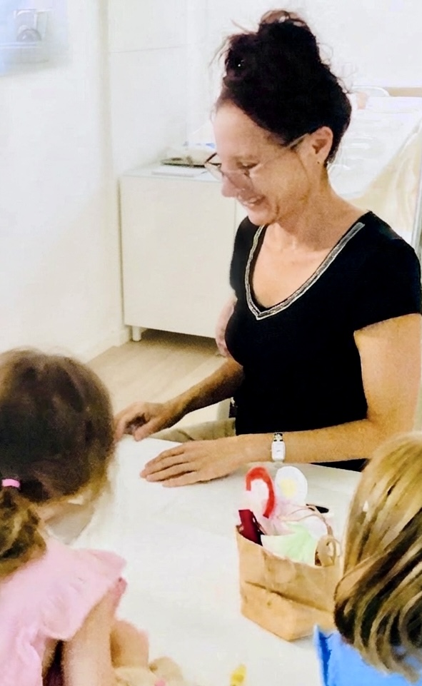 Andrea sitzt lachend mit zwei Kindern am Tisch, dazwischen ein Korb mit Papierfiguren